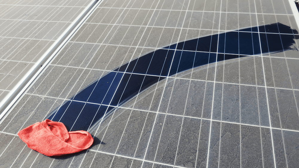 Image of a dusty solar panel being wiped with a cloth. The photo contrasts the difference between a clean and a dirty panel.